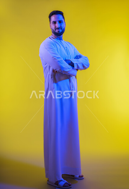 Standing straight with clasped hands, the concept of masculinity and self-confidence, a portrait of an Emirati Gulf Arab man wearing a kandura looking at the camera with an expression of pleasure, elegance and interest in his outward appearance, yellow background.