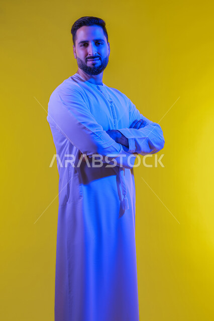 مفهوم الرجولة والثقة بالنفس, بورتريه لرجل عربي خليجي إماراتي يرتدي الكندورة ينظر الى الكاميرا بتعابير السرور، الوقوف باستقامة وايدي متشابكة ، الأناقة والاهتمام بالمظهر الخارجي، خلفية صفراء .