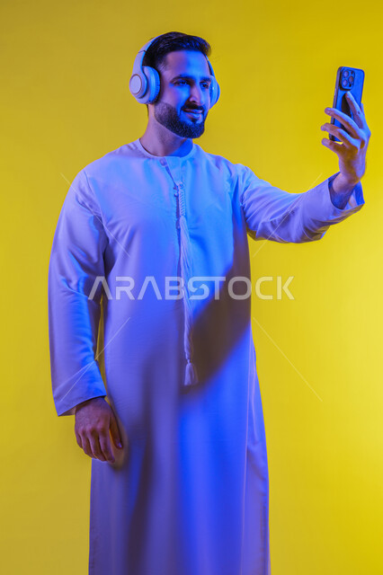عطلة نهاية الاسبوع ، الاستفادة من الهاتف المحمول والأجهزة التكنولوجية، شاب عربي خليجي اماراتى يرتدي الكندورة ويضع سماعة الرأس بإيماءات السعادة, استخدام تقنية الهيدفون، صورة الجسم بالطول الكامل, خلفية صفراء .