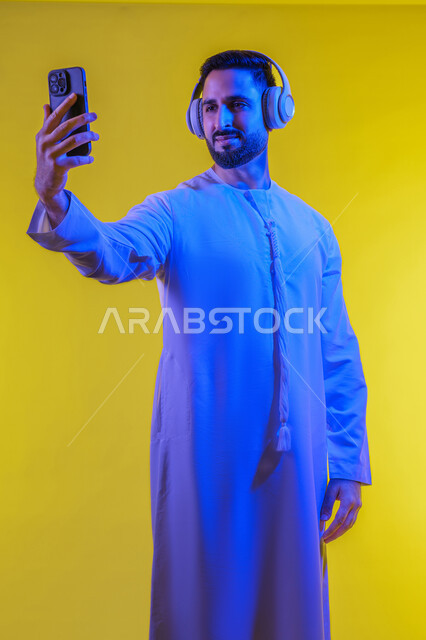 التقاط صور السيلفى ، الاستفادة من الهاتف المحمول والأجهزة التكنولوجية، شاب عربي خليجي اماراتى يرتدي الكندورة ويضع سماعة الرأس بإيماءات السعادة, استخدام تقنية الهيدفون، صورة الجسم بالطول الكامل, خلفية صفراء .