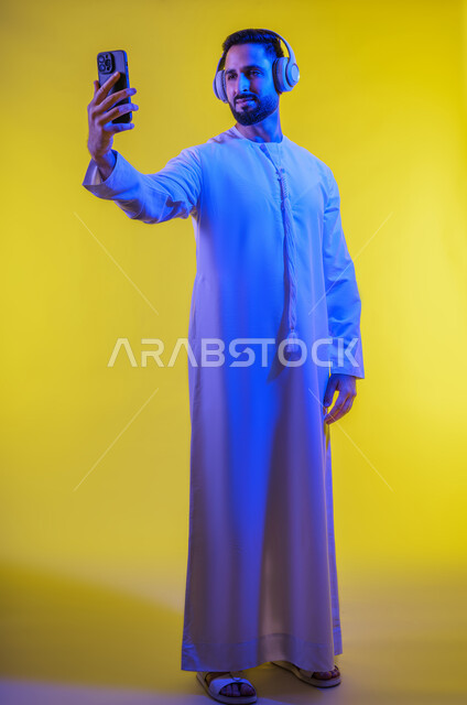 اجراء مكالمة فيديو عبر الجوال ، الاستفادة من الهاتف المحمول والأجهزة التكنولوجية، شاب عربي خليجي اماراتى يرتدي الكندورة ويضع سماعة الرأس بإيماءات السعادة, استخدام تقنية الهيدفون، صورة الجسم بالطول الكامل, خلفية صفراء .