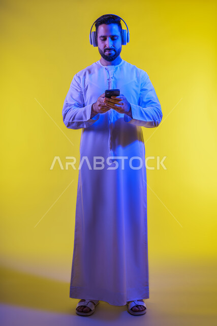 تطبيقات الاغاني والتسجيلات الصوتية في الهاتف المحمول، وضع سماعة الرأس ، مفهوم الإصغاء والاستماع، بورتريه من الجانب لرجل عربى خليجى اماراتى يرتدى الكندورة يقوم بإستخدام تقنية الهيدفون والجوال ، قضاء أوقات ممتعة بسماع الاغاني والمقاطع الصوتية، خلفية صفراء .