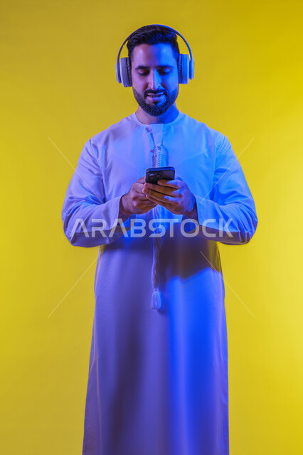 مفهوم الترفيه والتسلية في أوقات الفراغ، وضع سماعة الرأس ، مفهوم الإصغاء والاستماع، بورتريه من الجانب لرجل عربى خليجى اماراتى يرتدى الكندورة يقوم بإستخدام تقنية الهيدفون والجوال ، قضاء أوقات ممتعة بسماع الاغاني والمقاطع الصوتية، خلفية صفراء .