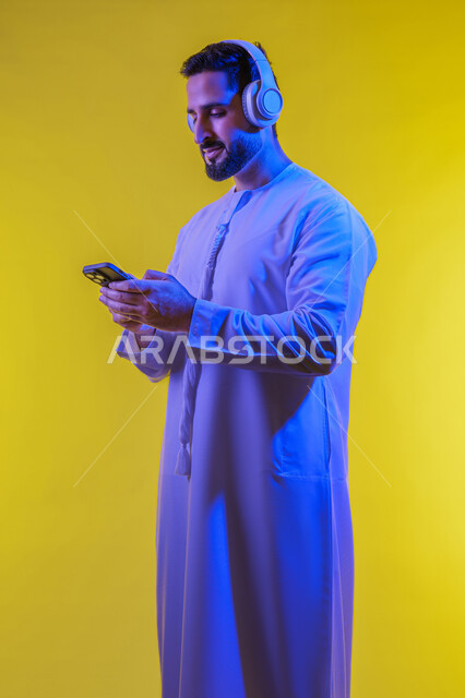 استخدام التقنيات الحديثة والمتطورة ، وضع سماعة الرأس ، مفهوم الإصغاء والاستماع، بورتريه من الجانب لرجل عربى خليجى اماراتى يرتدى الكندورة يقوم بإستخدام تقنية الهيدفون والجوال ، قضاء أوقات ممتعة بسماع الاغاني والمقاطع الصوتية، خلفية صفراء .