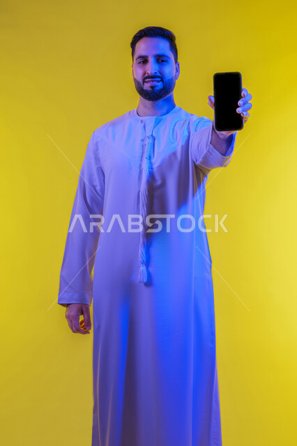 عرض تطبيقات وخدمات الهاتف المحمول ،عرض الجوال بشاشة سوداء فارغة، استخدام التكنولوجيا المتطورة للتسويق الرقمي الالكتروني ، بورتريه لشاب عربي خليجي اماراتى مبتسم يرتدي الكندورة ينظر إلى الكاميرا ،صورة الجسم بالطول الكامل، خلفية صفراء .