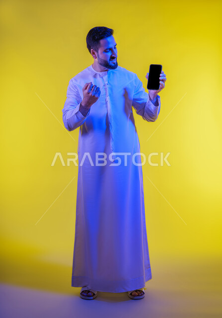 الحماس والدهشه بالفوز والنصر، عرض شاشة اعلانية سوداء فارغه على الجوال ، ايماءات الانفعال والاندماج أثناء مشاهدة مباراة كرة القدم، بورتريه لشاب عربي خليجي اماراتى يرتدي الكندورة ،صورة الجسم بالطول الكامل، خلفية صفراء .