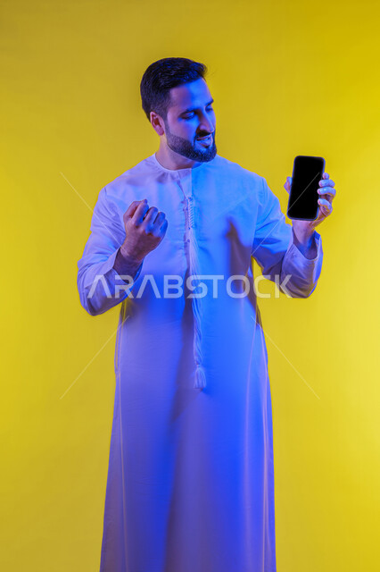 عرض شاشة اعلانية سوداء فارغه على الجوال، ايماءات الانفعال والاندماج أثناء مشاهدة مباراة كرة القدم، الحماس والدهشه بالفوز والنصر ، بورتريه لشاب عربي خليجي اماراتى يرتدي الكندورة ،صورة الجسم بالطول الكامل، خلفية صفراء .