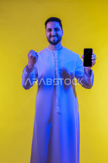 ايماءات الانفعال والاندماج أثناء مشاهدة مباراة كرة القدم، عرض شاشة اعلانية سوداء فارغه على الجوال، الحماس والدهشه بالفوز والنصر ، بورتريه لشاب عربي خليجي اماراتى يرتدي الكندورة ينظر إلى الكاميرا ،صورة الجسم بالطول الكامل، خلفية صفراء .