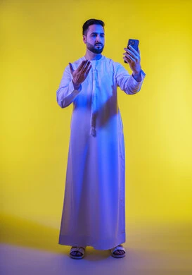 Live video broadcast, social media use, portrait of an Emirati Gulf Arab man wearing the Emirati kandura, looking at his mobile phone in surprise and astonishment, facial and hand gestures indicating surprise, amazement and amazement, yellow background.