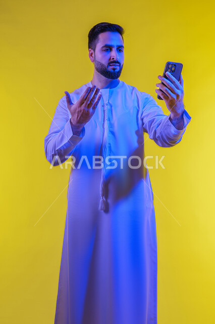 إيماءات وجه ويد تدل على الدهشة والذهول والإنبهار، بورتريه لرجل عربي خليجي إماراتي يرتدي الكندورة الإماراتية ويقوم بالنظر إلى الهاتف المحمول بتعجب وإستغراب، بث فيديو مباشر، إستخدام وسائل التواصل الإجتماعي، خلفية صفراء .