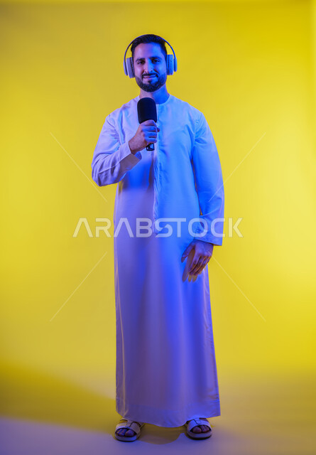 الاستماع للأغاني والمواهب الغنائية، التفاعل والاندماج مع الأنغام الموسيقية, إيماءات الإندماج والتفاعل مع الغناء، بورتريه لرجل عربي خليجي إماراتي يرتدي الكندورة الإماراتية ، يضع سماعات الرأس ويمسك المايكروفون ، تسجيل مقطع صوتي غنائي، خلفية صفراء .