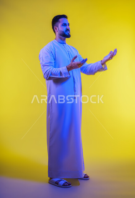 محاكاة العالم الخيالى ، مفهوم الاناقة و الاهتمام بالمظهر الخارجي, إحتواء وإمساك عنصر ما، بورتريه لرجل عربي خليجي اماراتي يرتدي الكندورة يفتح يديه لعرض عناصر هولوجرامية ثلاثية الأبعاد، صورة الجسم بالطول الكامل, خلفية صفراء .