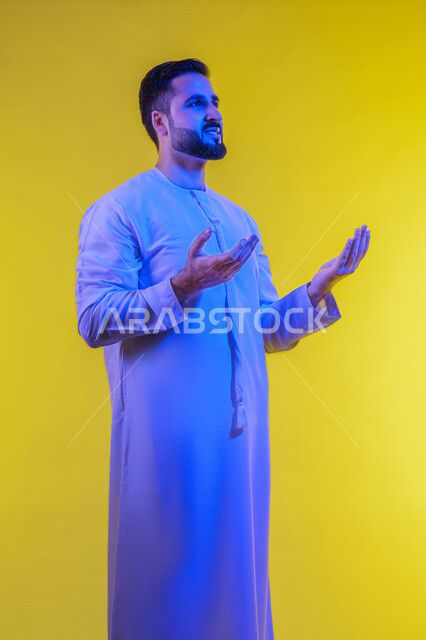 تعابير السرور والسعادة, مفهوم الاناقة و الاهتمام بالمظهر الخارجي, إحتواء وإمساك عنصر ما، بورتريه لرجل عربي خليجي اماراتي يرتدي الكندورة يفتح يديه لعرض عناصر هولوجرامية ثلاثية الأبعاد، صورة الجسم بالطول الكامل, خلفية صفراء .