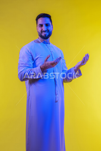 رفع شيء معين, تعابير حمل و إمساك عنصر محدد ، مفهوم الاناقة و الاهتمام بالمظهر الخارجي, بورتريه لشاب عربي خليجي اماراتي يرتدي الكندورة يفتح يديه لعرض عناصر هولوجرامية ثلاثية الأبعاد، صورة الجسم بالطول الكامل, خلفية صفراء .