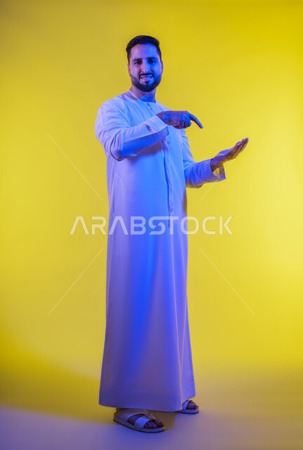 مفهوم الاناقة و الاهتمام بالمظهر الخارجي, إحتواء وإمساك عنصر ما،  بورتريه لشاب عربي خليجي اماراتي يرتدي الكندورة يفتح يديه لعرض عناصر هولوجرامية ثلاثية الأبعاد، صورة الجسم بالطول الكامل, خلفية صفراء .