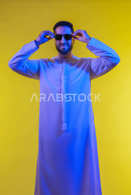 استخدام العدسات الطبية لحماية العين من أشعة الشمس، الأناقة والوسامة والاهتمام بالمظهر الخارجي، بورتريه لرجل عربي خليجي اماراتي يرتدي الكندورة ويضع نظارة شمسية سوداء، إيماءات الثقة بالنفس ، الجسم بالطول الكامل, خلفية صفراء .