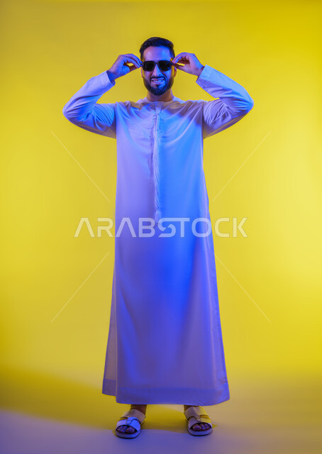 الأناقة والوسامة والاهتمام بالمظهر الخارجي، استخدام العدسات الطبية لحماية العين من أشعة الشمس، بورتريه لرجل عربي خليجي اماراتي يرتدي الكندورة ويضع نظارة شمسية سوداء، إيماءات الثقة بالنفس ، الجسم بالطول الكامل, خلفية صفراء .