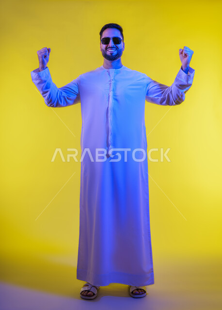 التمتع بالبنية الجسدية القوية، بورتريه لرجل عربي خليجي إماراتي يرتدي الكندورة الإماراتية ونظارات الشمس ، يقوم بعرض عضلات يديه، إيماءات وجه ويدين تدل على القوة العضلية، اللياقة البدنية والجسم الصحي القوى، خلفية صفراء .