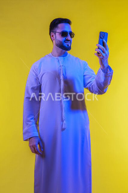 دمج التقنيات والتكنولوجيا الحديثة بالحياة اليومية، اجراء مكالمة فيديو والتواصل مع الاهل والاصدقاء، التقاط صور السيلفي باستخدام الهاتف المحمول، توثيق اللقطات الشخصية التذكارية، بورتريه لرجل عربي خليجي اماراتي يرتدي الكندورة يمسك الجوال، خلفية صفراء .
