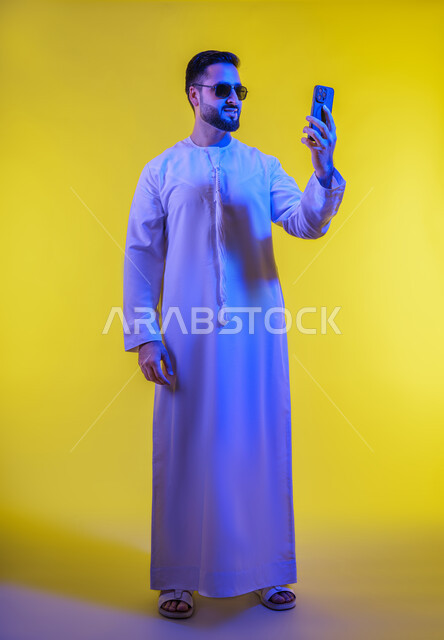 توثيق اللقطات الشخصية التذكارية، التقاط صور السيلفي باستخدام الهاتف المحمول، دمج التقنيات والتكنولوجيا الحديثة بالحياة اليومية، اجراء مكالمة فيديو والتواصل مع الاهل والاصدقاء، بورتريه لرجل عربي خليجي اماراتي يرتدي الكندورة يمسك الجوال، خلفية صفراء .