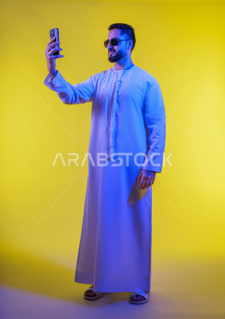 Taking selfies with a mobile phone, integrating modern technology into daily life, making a video call and communicating with family and friends, documenting personal memories, a portrait of an Emirati Gulf Arab man wearing a kandura holding a mobile phone, yellow background.