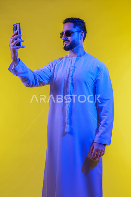 اجراء مكالمة فيديو والتواصل مع الاهل والاصدقاء، التقاط صور السيلفي باستخدام الهاتف المحمول، دمج التقنيات والتكنولوجيا الحديثة بالحياة اليومية، توثيق اللقطات الشخصية التذكارية، بورتريه لرجل عربي خليجي اماراتي يرتدي الكندورة يمسك الجوال، خلفية صفراء .
