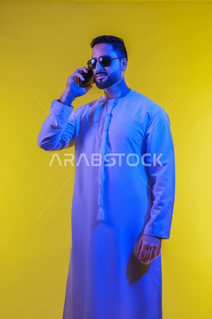 استخدام التكنولوجيا والأجهزة التقنية الحديثة في الحياة اليومية، دردشة ومحادثات والتواصل مع الأفراد ، إجراء مكالمات هاتفية باستخدام الهاتف المحمول , بورتريه لرجل عربي خليجي اماراتى يرتدي الكندورة والنضارة الشمسية ويستخدم الجوال ، صورة الجسم بالطول الكامل , خلفية صفراء .