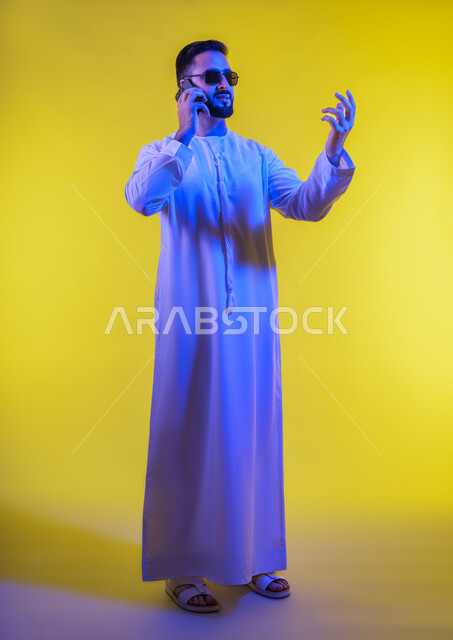 Answering calls and messages, using technology and modern devices in daily life. Portrait of an Emirati Gulf Arab man wearing a kandura and sunglasses, using a mobile phone. Making phone calls using a mobile phone. Full-length body image. Yellow background.