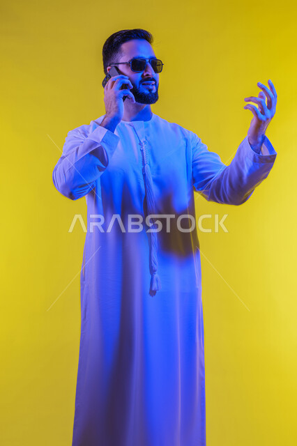 Facial expressions indicating question, answering calls and messages, portrait of an Arab Gulf Emirati man wearing a kandura and sunglasses and using a mobile phone, making phone calls using a mobile phone, full body image, yellow background.