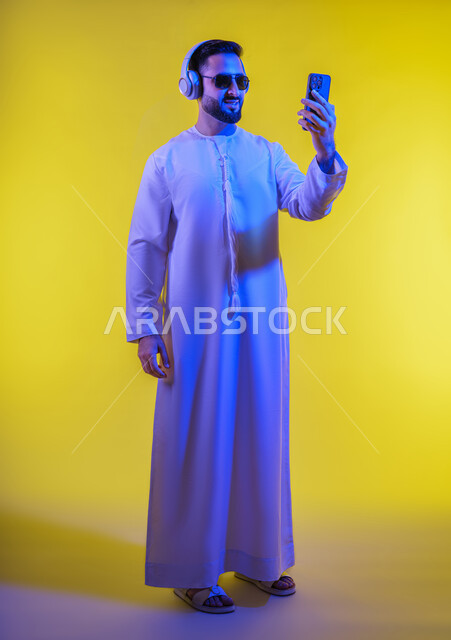 اجراء بث مباشر ، استخدام تقنية الهيدفون، الاستفادة من الجوال والأجهزة التكنولوجية، شاب عربي خليجي اماراتى يرتدي الكندورة ويضع سماعة الرأس بإيماءات السعادة,  الاستماع إلى الموسيقى والتسجيلات الصوتية، صورة الجسم بالطول الكامل, خلفية صفراء .