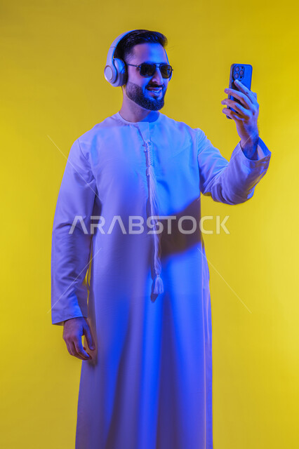 استخدام تقنية الهيدفون، الاستفادة من الجوال والأجهزة التكنولوجية، شاب عربي خليجي اماراتى يرتدي الكندورة ويضع سماعة الرأس بإيماءات السعادة,  الاستماع إلى الموسيقى والتسجيلات الصوتية، صورة الجسم بالطول الكامل, خلفية صفراء .