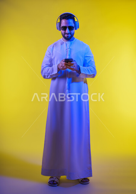 الاستماع إلى الموسيقى والتسجيلات الصوتية، الاستفادة من الجوال والأجهزة التكنولوجية، شاب عربي خليجي اماراتى يرتدي الكندورة ويضع سماعة الرأس بإيماءات السعادة, استخدام تقنية الهيدفون، صورة الجسم بالطول الكامل, خلفية صفراء .
