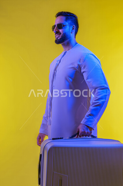 Escaping from life's pressures, preparing for an enjoyable trip, going on recreational tours during the summer vacation, having a good time outside the country, tourist activities and events, expressions of joy and happiness, a side portrait of a smiling young Arab Gulf Emirati man wearing a kandura and sunglasses, holding a travel bag, yellow background.