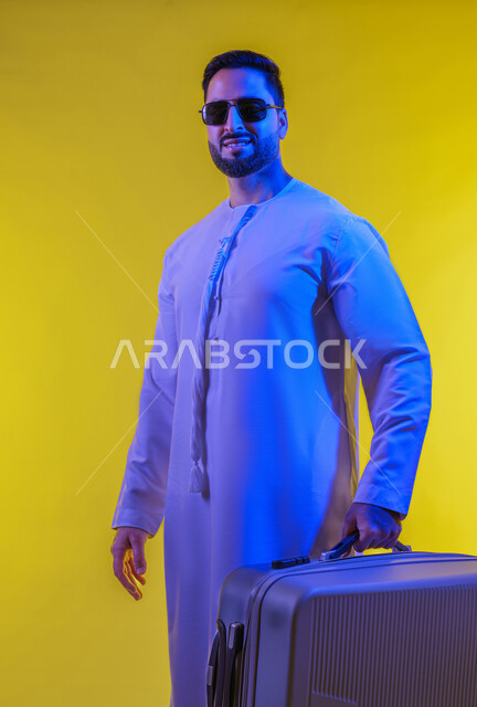 الاستعداد للسفر الممتع, الهروب من ضغوط الحياة، الذهاب في جولات ترفيهية في إجازة الصيف, قضاء أوقات ممتعه خارج البلاد, نشاطات وفعاليات سياحية, تعابير الفرح والسرور، بورتريه من الجانب لشاب عربي خليجي اماراتى مبتسم يرتدي الكندورة والنظارة الشمسية  يمسك بيده حقيبة السفر، خلفية صفراء .