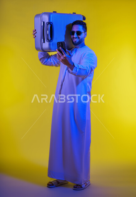 إجراء مكالمات فيديو عبر الجوال, التواصل مع الأهل والأصدقاء,  بورتريه لشاب عربي اماراتى خليجي مبتسم يرتدي الكندورة الاماراتية يمسك بيده الهاتف المحمول ويحمل حقيبة السفر, حجز مباشر عبر تطبيقات وبرامج السياحة, صورة الجسم بالطول الكامل, خلفية صفراء .