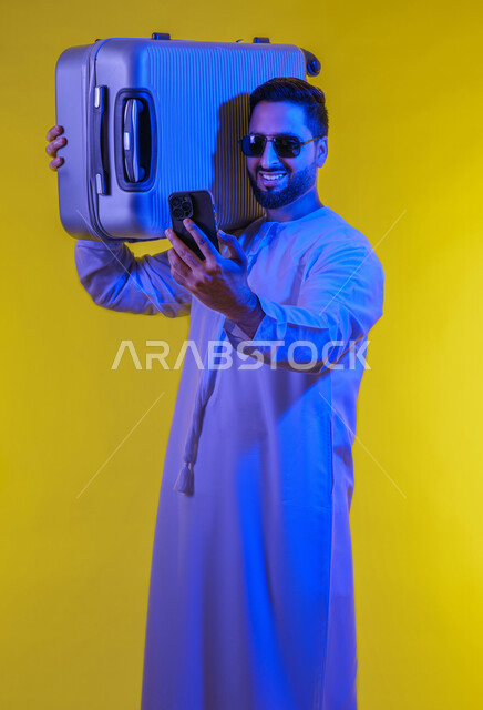 التواصل مع الأهل والأصدقاء, إجراء مكالمات فيديوعبر الجوال, بورتريه لشاب عربي اماراتى خليجي مبتسم يرتدي الكندورة الاماراتية يمسك بيده الهاتف المحمول ويحمل حقيبة السفر, حجز مباشر عبر تطبيقات وبرامج السياحة, صورة الجسم بالطول الكامل, خلفية صفراء .
