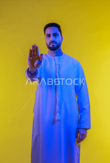 مفهوم الرفض والامتناع ، ايماءات طلب التوقف وعدم الاقتراب، بورتريه لرجل عربي خليجي اماراتى يرتدي الكندورة يقف باستقامة يرفع كفيه للاعلى، النظر الى الكاميرا بتعابير الحزم، الجسم الكامل بالطول، خلفية صفراء .