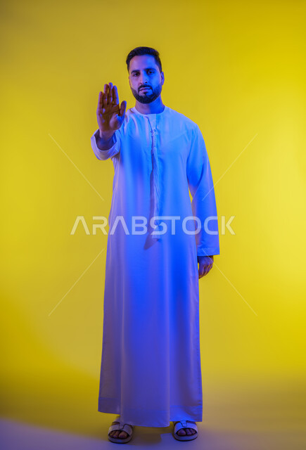 Looking at the camera with a firm expression, the concept of refusal and abstention, gestures asking to stop and not approach, a portrait of an Arab Gulf Emirati man wearing a kandura, standing straight with his hands raised, full body length, yellow background.