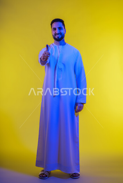 الامتياز والعلامة الكاملة، التعبير عن الإعجاب والتقدير، رفع اليد بعلامات الجودة والقبول، الشعور بالرضا والاستحسان، بورتريه لرجل عربي خليجي اماراتي يرتدي الكندورة ينظر إلى الكاميرا بإيماءات السعادة والسرور، الجسم بالطول الكامل، خلفية صفراء .