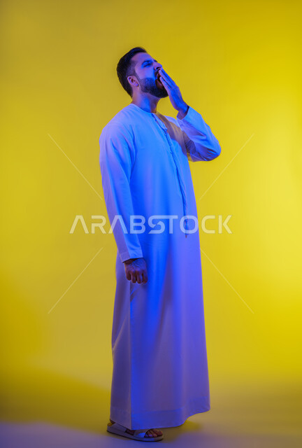 إيماءات التعب والإرهاق، ضرورة أخذ قسط من الراحة والاسترخاء، الشعور بالنعاس والحاجة للنوم، بورتريه لرجل عربي خليجي اماراتي يرتدي الكندورة يقوم بوضع يده أمام فمه والتثاؤب، خلفية صفراء .