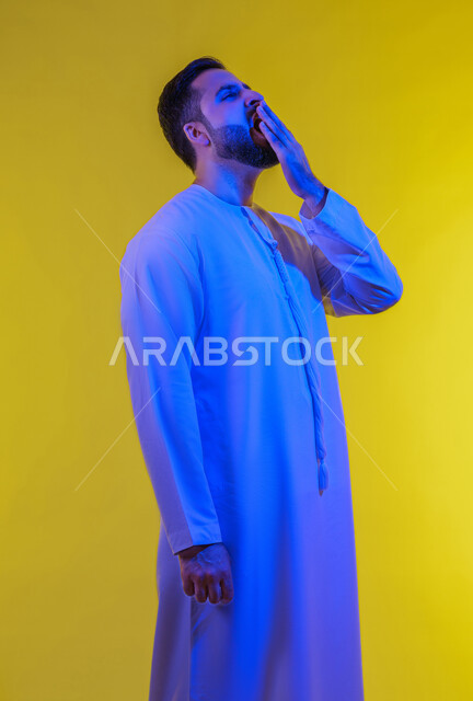 الشعور بالنعاس والحاجة للنوم، ضرورة أخذ قسط من الراحة والاسترخاء، إيماءات التعب والإرهاق، بورتريه لرجل عربي خليجي اماراتي يرتدي الكندورة يقوم بوضع يده أمام فمه والتثاؤب، خلفية صفراء .