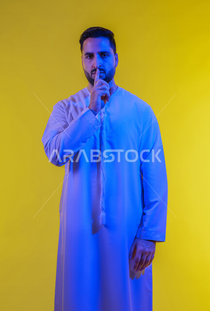 طلب التوقف عن الكلام والصمت، النهي والزجر والانصات، وضع اصبع السبابة أمام الفم بإيماءات الجدية، الأمر بالصمت التام، بورتريه لرجل عربي خليجي اماراتي يرتدي الكندورة  ينظر للكاميرا بإيماءات الغضب، الجسم بالطول الكامل، خلفية صفراء.