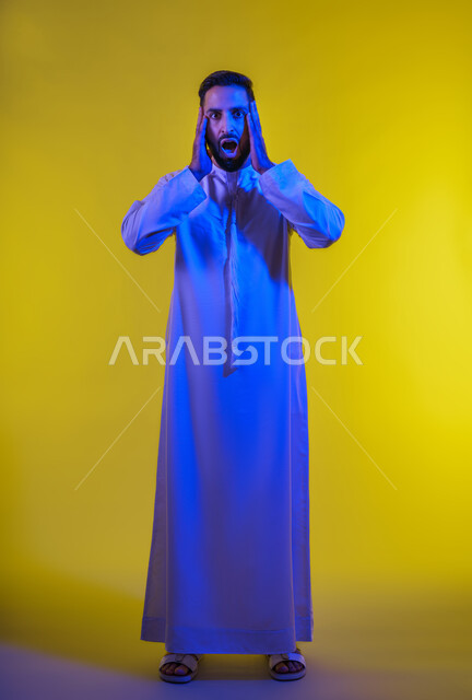 النظر الى الكاميرا بتعابير التعجب, الانزعاج عند سماع خبر سيء ,الوقوع في مصيبة, الصدمة و المفاجأة برؤية شيء ما، بورتريه لرجل عربي اماراتي خليجي يرتدي الكندورة يضع يديه على رأسه بتعابير الدهشة و الذهول، صورة الجسم بالطول الكامل، خلفية صفراء .