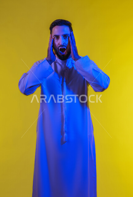 الانزعاج عند سماع خبر سيء ,النظر الى الكاميرا بتعابير التعجب, الوقوع في مصيبة, الصدمة و المفاجأة برؤية شيء ما، بورتريه لرجل عربي اماراتي خليجي يرتدي الكندورة يضع يديه على رأسه بتعابير الدهشة و الذهول، صورة الجسم بالطول الكامل، خلفية صفراء .