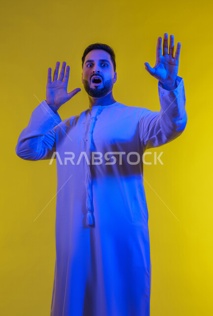 Stop, refrain and refrain from approaching, self-protection from external harm, portrait of an Arab Gulf Emirati man wearing a kandura looking at the camera and raising his hands with an expression of fear and distancing, yellow background.