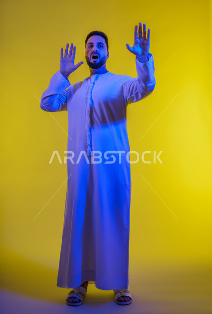 حماية النفس من ضرر خارجي ، طلب التوقف و الامتناع و عدم الاقتراب, بورتريه لرجل عربي خليجي إماراتي يرتدي الكندورة ينظر إلى الكاميرا و يرفع يديه بتعابير الخوف و الابتعاد, خلفية صفراء.