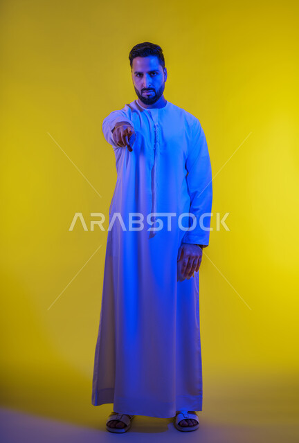 التحديد نحو هدف معين, ايماءات التأكد من شيء ما , بورتريه لرجل عربي اماراتي خليجي يرتدي الكندورة يشير الى الكاميرا باصبع السبابة , صورة الجسم بالطول الكامل, خلفية صفراء.