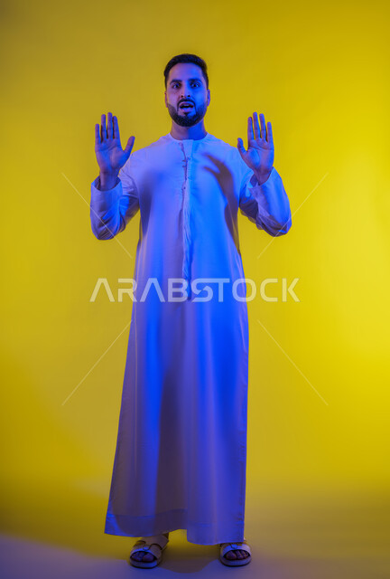 ايماءات طلب التوقف وعدم الاقتراب، مفهوم الرفض والامتناع ، بورتريه لرجل عربي خليجي اماراتى يرتدي الكندورة يقف باستقامة يرفع كفيه للاعلى، عرض باطن الكف والنظر الى الكاميرا بتعابيرالحزم، الجسم الكامل بالطول، خلفية صفراء .
