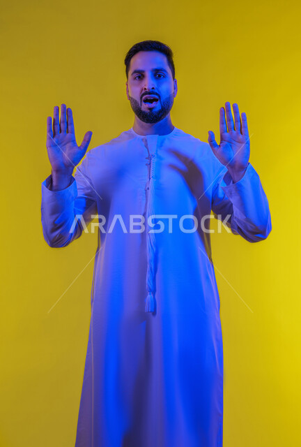 مفهوم الرفض والامتناع ,ايماءات طلب التوقف وعدم الاقتراب، بورتريه لرجل عربي خليجي اماراتى يرتدي الكندورة يقف باستقامة يرفع كفيه للاعلى، عرض باطن الكف والنظر الى الكاميرا بتعابيرالحزم، الجسم الكامل بالطول، خلفية صفراء .