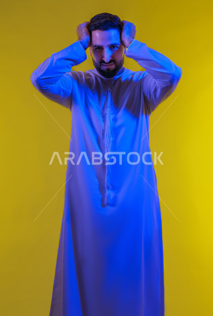 المفاجأة و الصدمة عند رؤية شيء ما، الانزعاج عند سماع خبر سيء ,الوقوع في مصيبة, النظر الى الكاميرا بتعابير التعجب, بورتريه لرجل عربي اماراتي خليجي يرتدي الكندورة يضع يديه على رأسه بتعابير الدهشة و الذهول، خلفية صفراء .